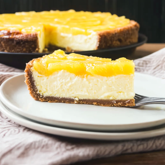 Slice of creamy cheesecake topped with a layer of yellow fruit on a white plate, with the rest of the cheesecake in the background.